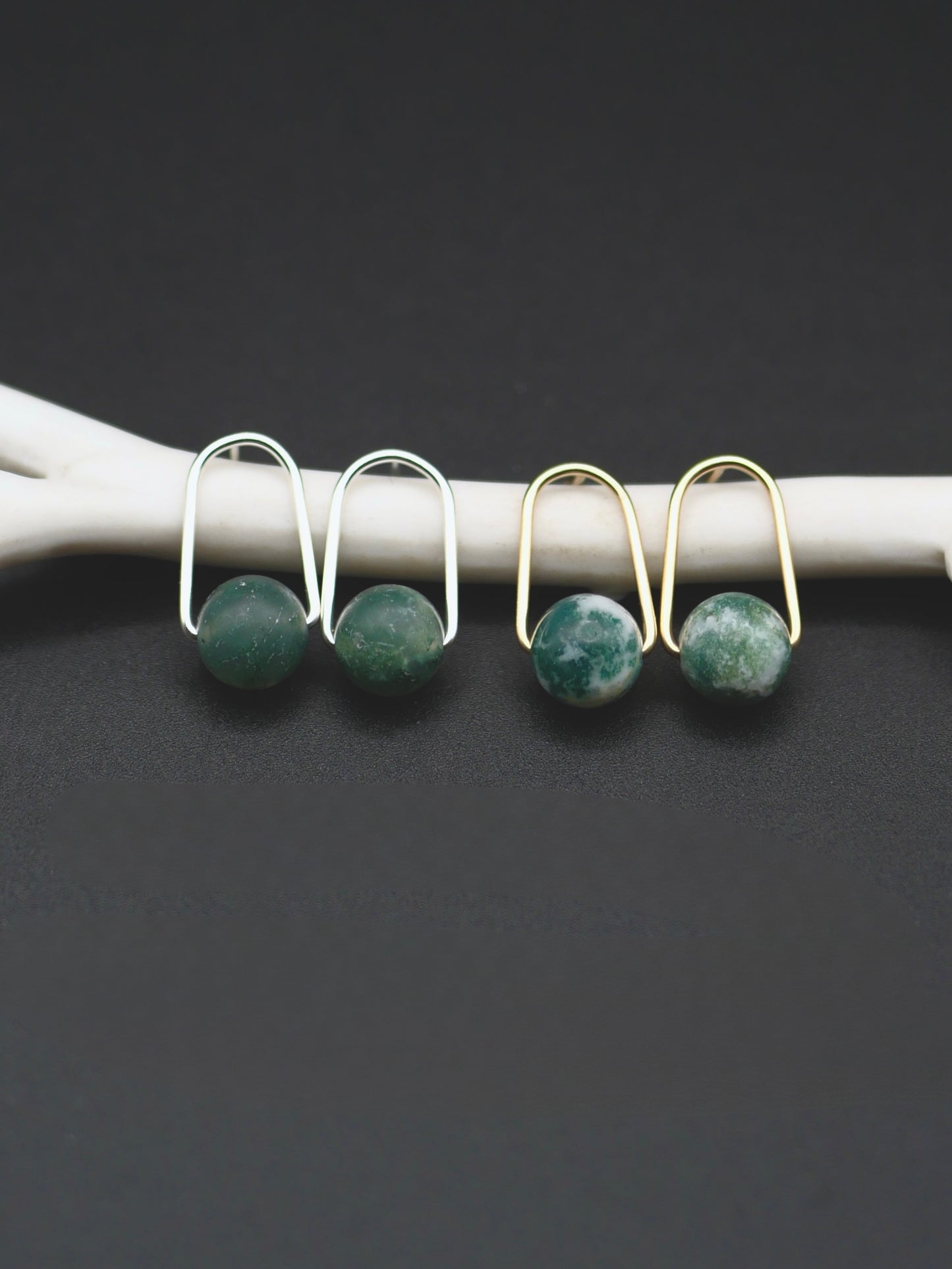 Two pairs of earrings with round green stone drops, shown on a white curved display against a dark gray background. One pair is silver-toned and one is gold-toned.