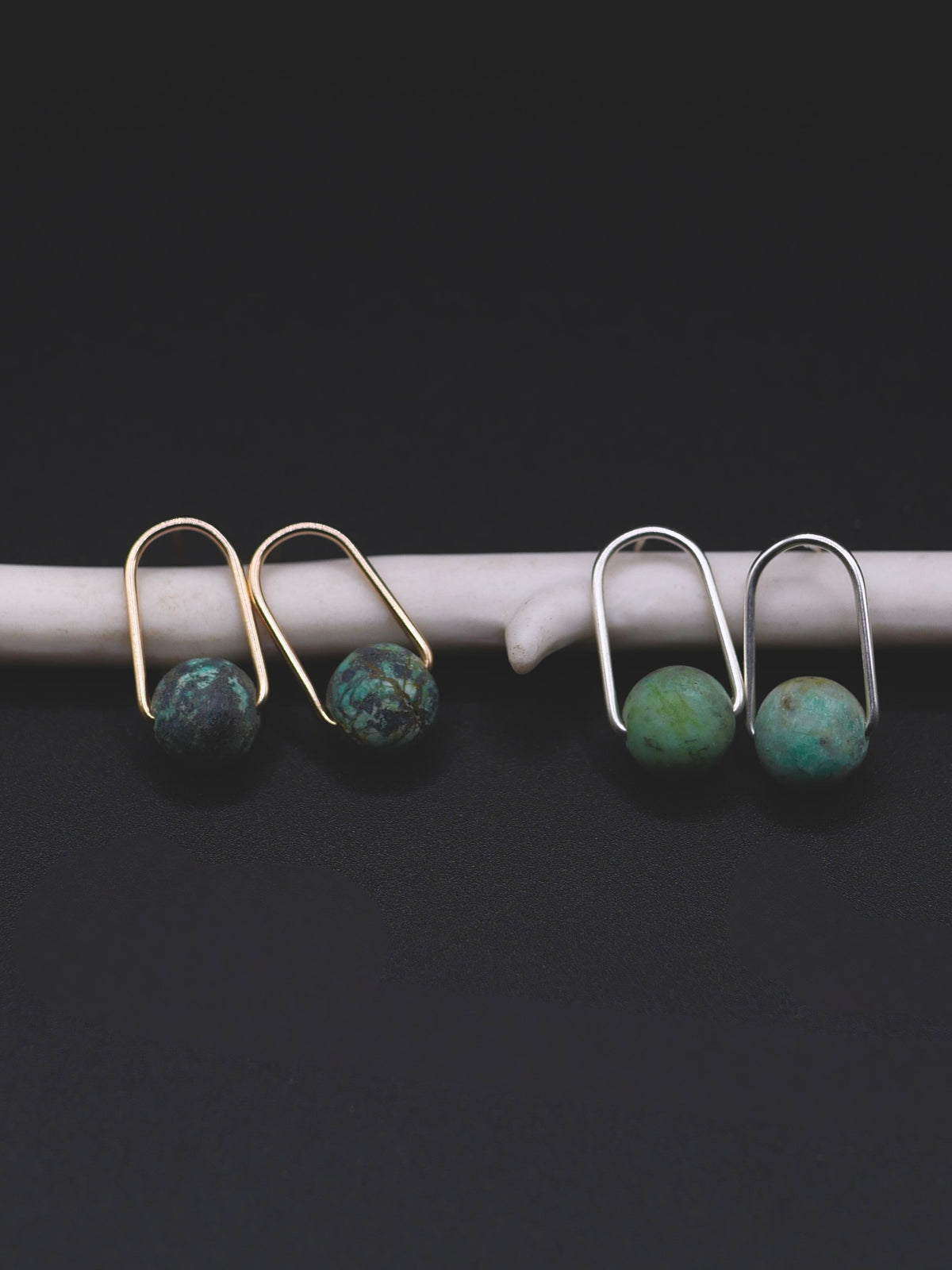 Small stud earrings with round turquoise-blue beads, shown in two pairs: one with gold-toned metal and one with silver-toned metal, resting on a white curved display against a black background.