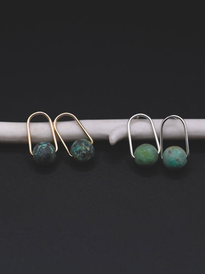 Small stud earrings with round turquoise-blue beads, shown in two pairs: one with gold-toned metal and one with silver-toned metal, resting on a white curved display against a black background.