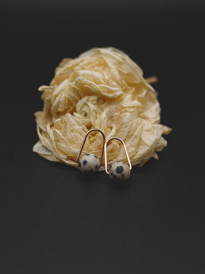 A pair of small gold-toned hoop earrings with round black-and-white spotted beads, displayed on a flower against a dark background.
