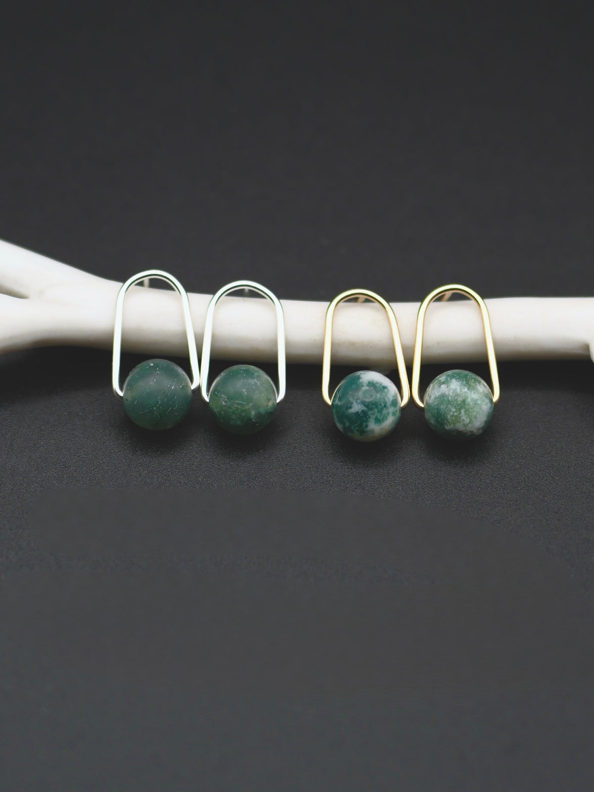 Two pairs of earrings with round green stone drops, shown on a white curved display against a dark gray background. One pair is silver-toned and one is gold-toned.
