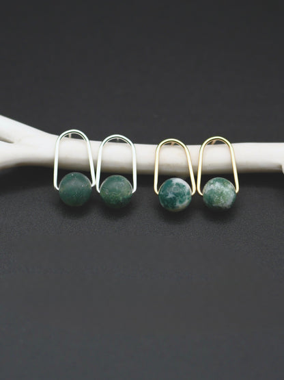 Two pairs of earrings with round green stone drops, shown on a white curved display against a dark gray background. One pair is silver-toned and one is gold-toned.
