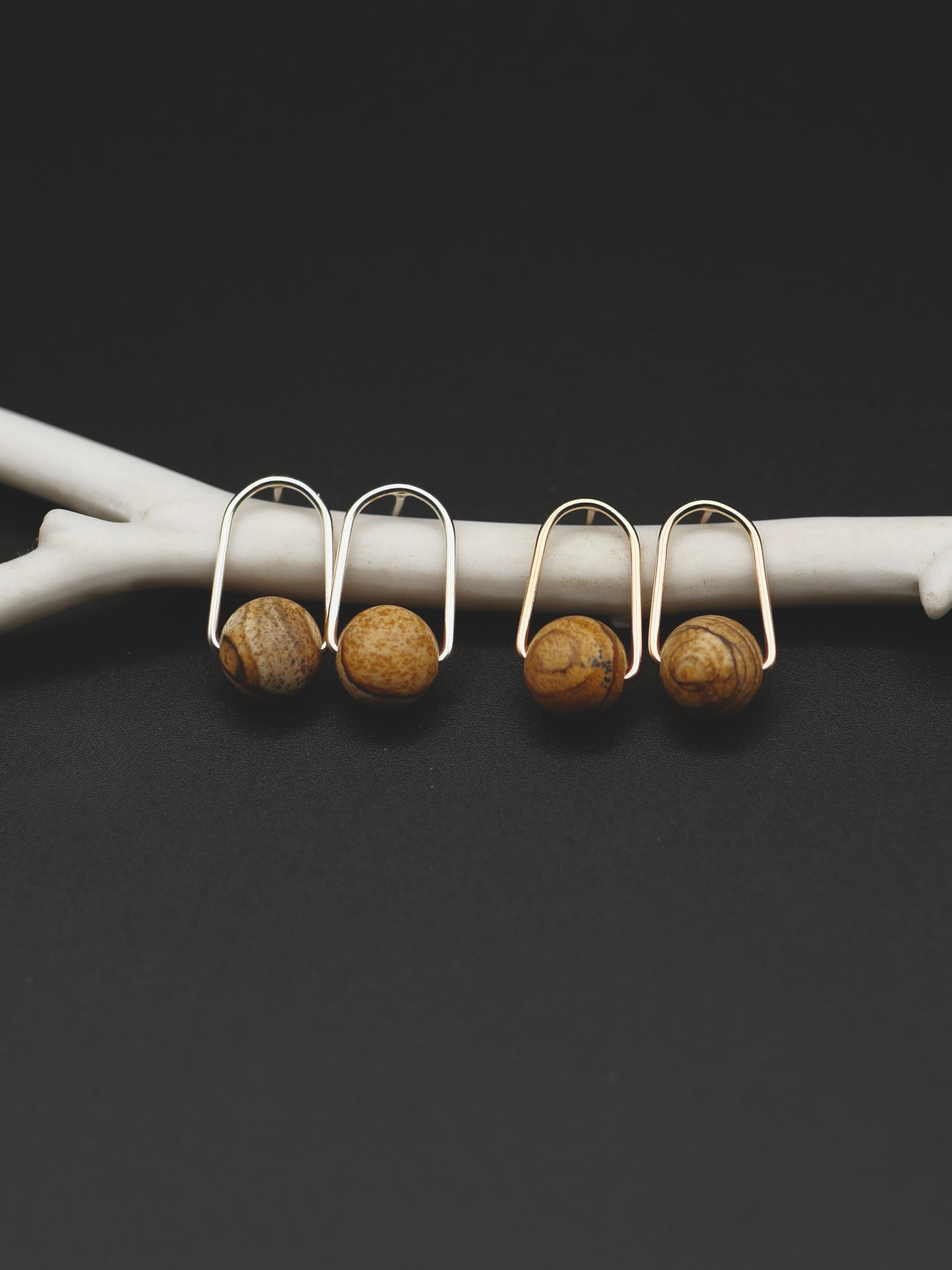 Two pairs of small earrings with round brown jasper beads, displayed on a white branch against a dark background.