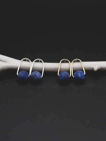 Two pairs of small hoop earrings with round sodalite stones, shown hanging from a white branch against a dark background. One pair is silver-toned and one pair is gold-toned.