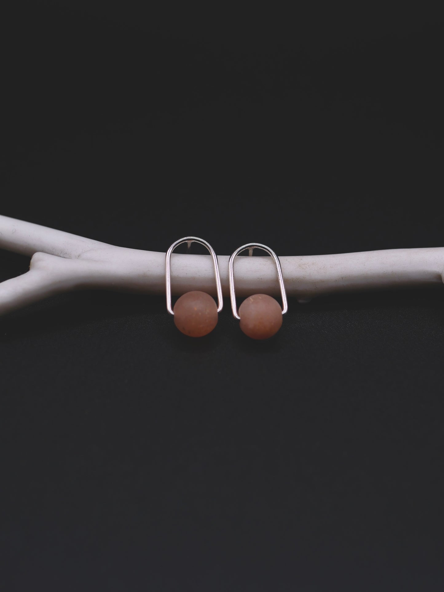 A pair of minimalist silver-toned drop earrings with smooth, matte light sunstone  beads, displayed on a light gray branch against a dark background.