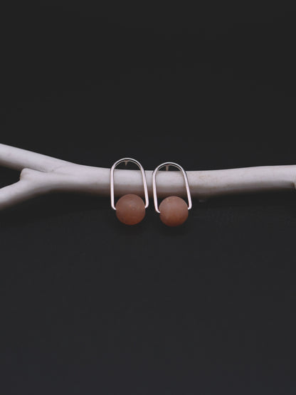 A pair of minimalist silver-toned drop earrings with smooth, matte light sunstone  beads, displayed on a light gray branch against a dark background.