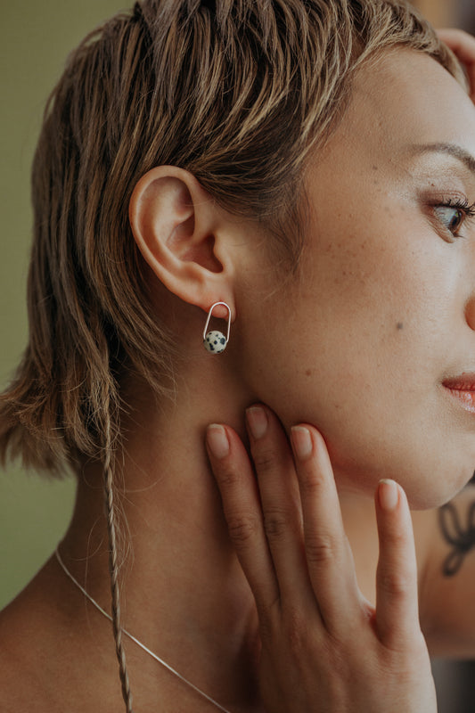 A close-up lifestyle shot of a model wearing a small silver stud earring with a gemstone. The model has short, textured blonde hair and is touching her neck, with a soft olive green background.